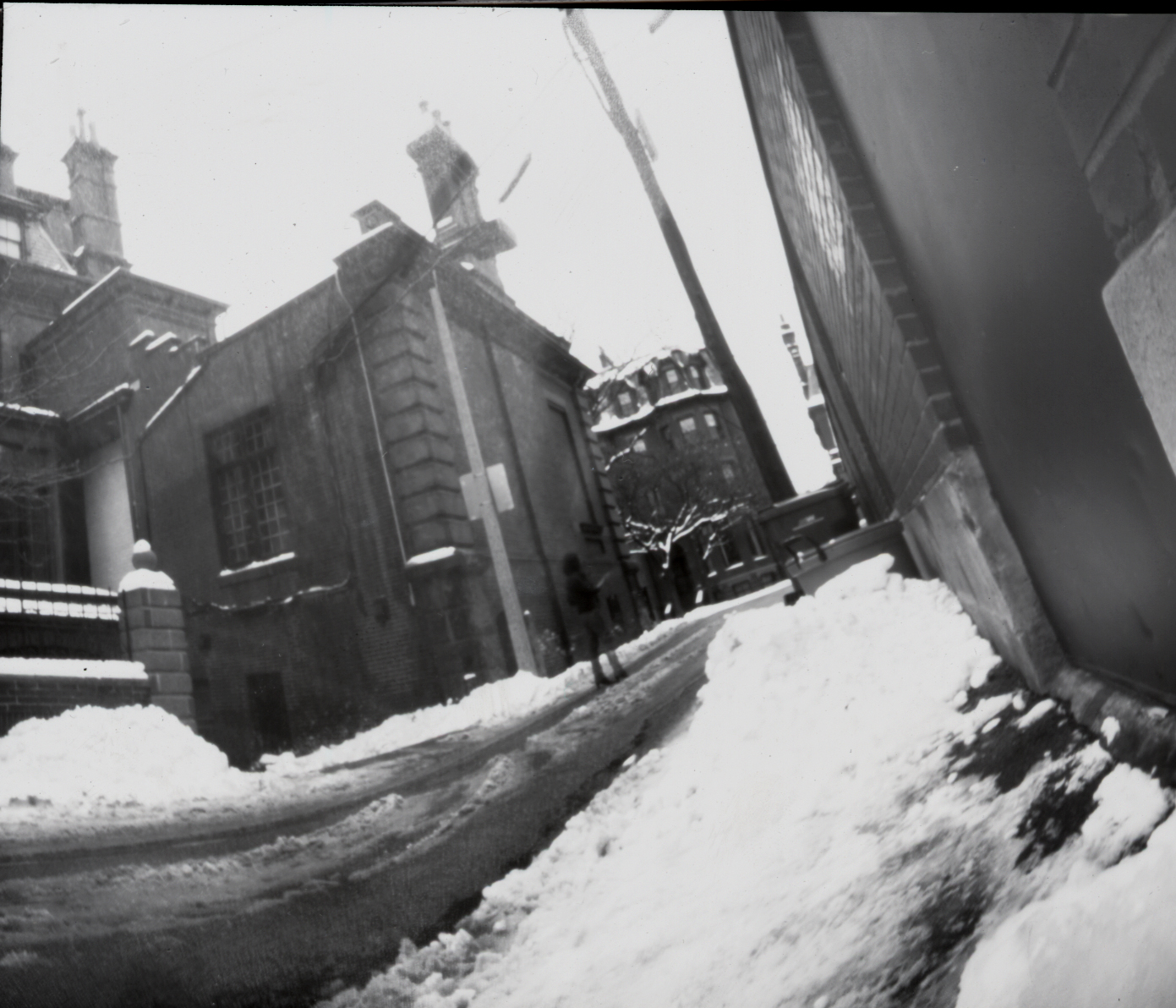 a black and white pinhole camera photograph with a large amount of curvature distortion except at the center of the image; the photo is shot at a diagonal angle on the ground and shows the alley behind a brownstone building in back bay in winter; the alley is lined with dirty snow; a figure in the center of the image stands far away from the camera and facing away