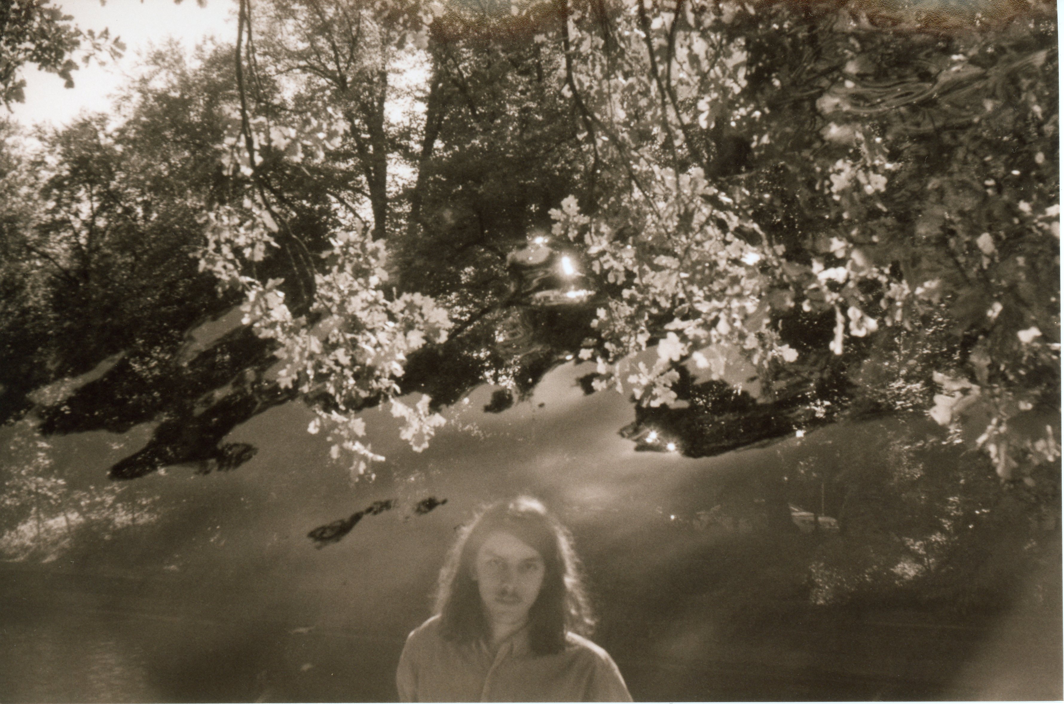 a black and white, sepia toned, 35mm print; the image is a hectic double exposure: one image is the surface of the water with faint ripples and sunlight on the surface reflected through trees; the other image shows a man looking at the camera in the center of the frame at the bottom, his head and shoulders visible; he stands in front of a canal with trees growing behind it
