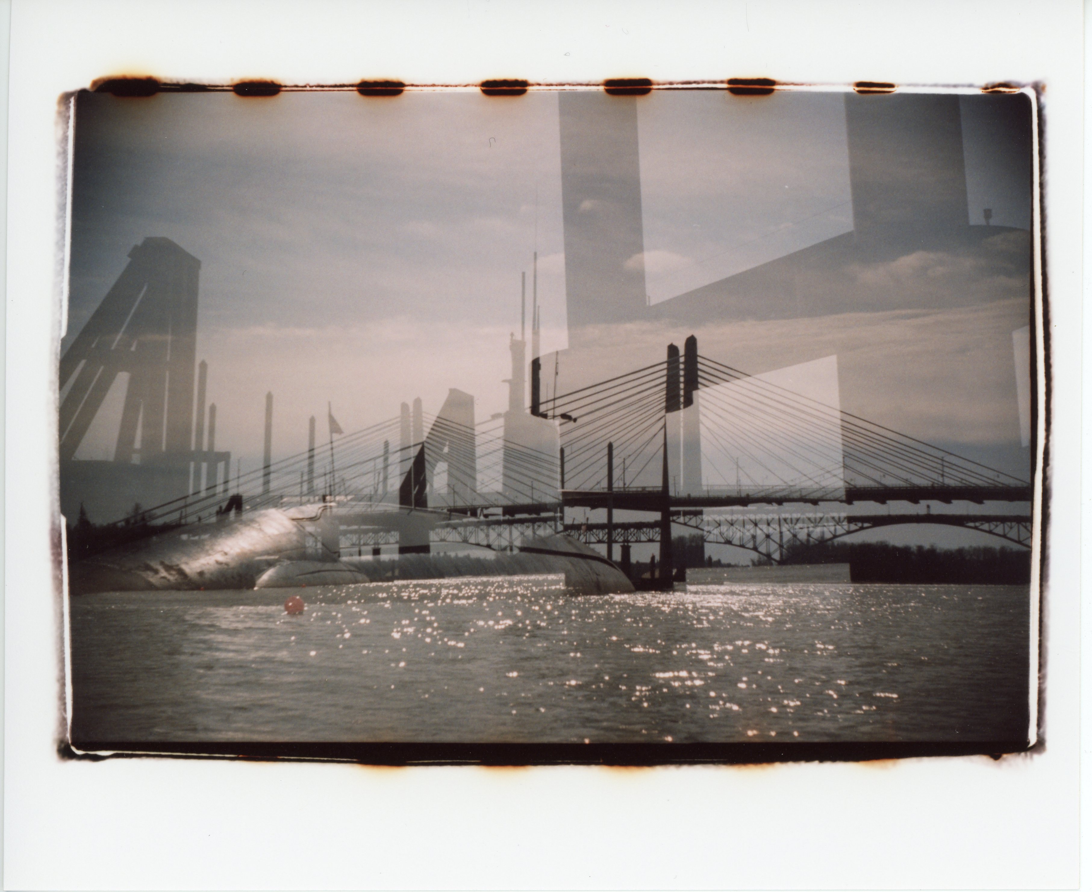 a 35mm color print with messy edges that show bits of the sprocket holes; the lower third of the image is water sparkling in the sun with a small red buoy floating a ways off towards the left side; the middle third shows the pilings, arches, and suspension cables of multiple exposures of docks and bridges; the upper third is mostly sky which is sunny with some clouds and hazy; on the right of the image, faintly in the foreground are two large pilings connected by a beam; on the left side the shiny round hull of a submarine that is part of OMSI is partially visible