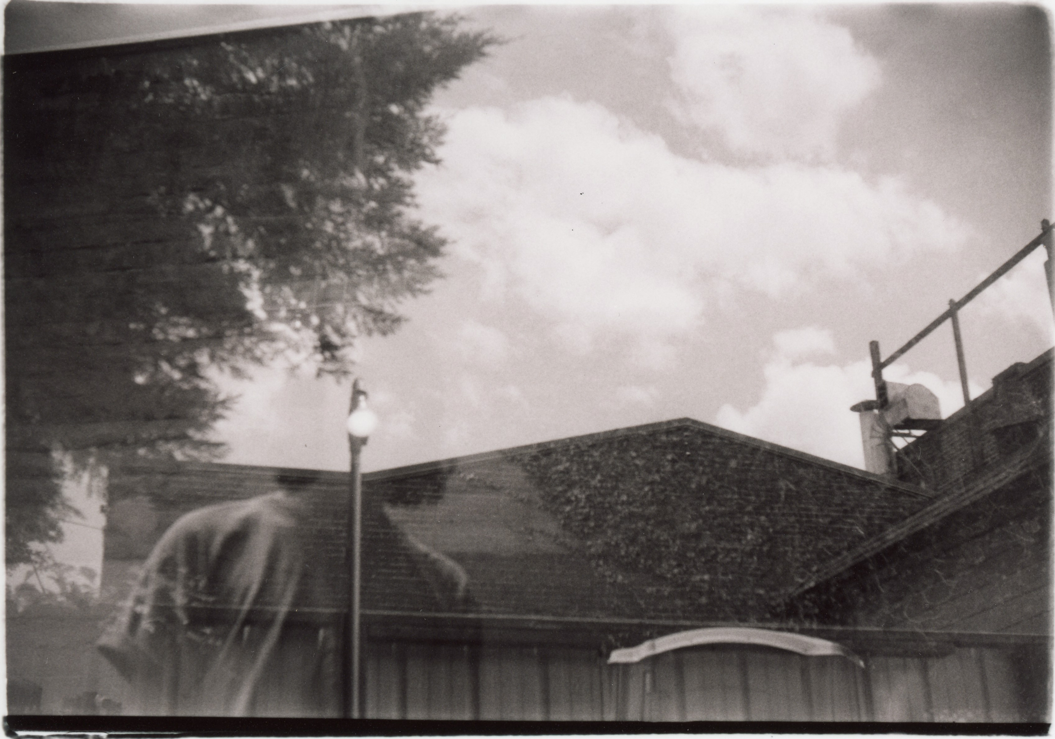 a black and white, sepia toned, 35mm print with messy edges; the image is a double exposure; one image shows a lamp pole, the lamp is on during a partially cloudy day; the lamp pole stands in front of a strip of corrugated metal at the bottom of the frame, which is in front of a shingled roofline; there is a tree in the top left corner, and on the right some ducts bars come out of the roof; in the second image a person is standing overlaid with the lamp pole; the light shines where their face would be, but the sky from the other image obscures the exposure of their head
