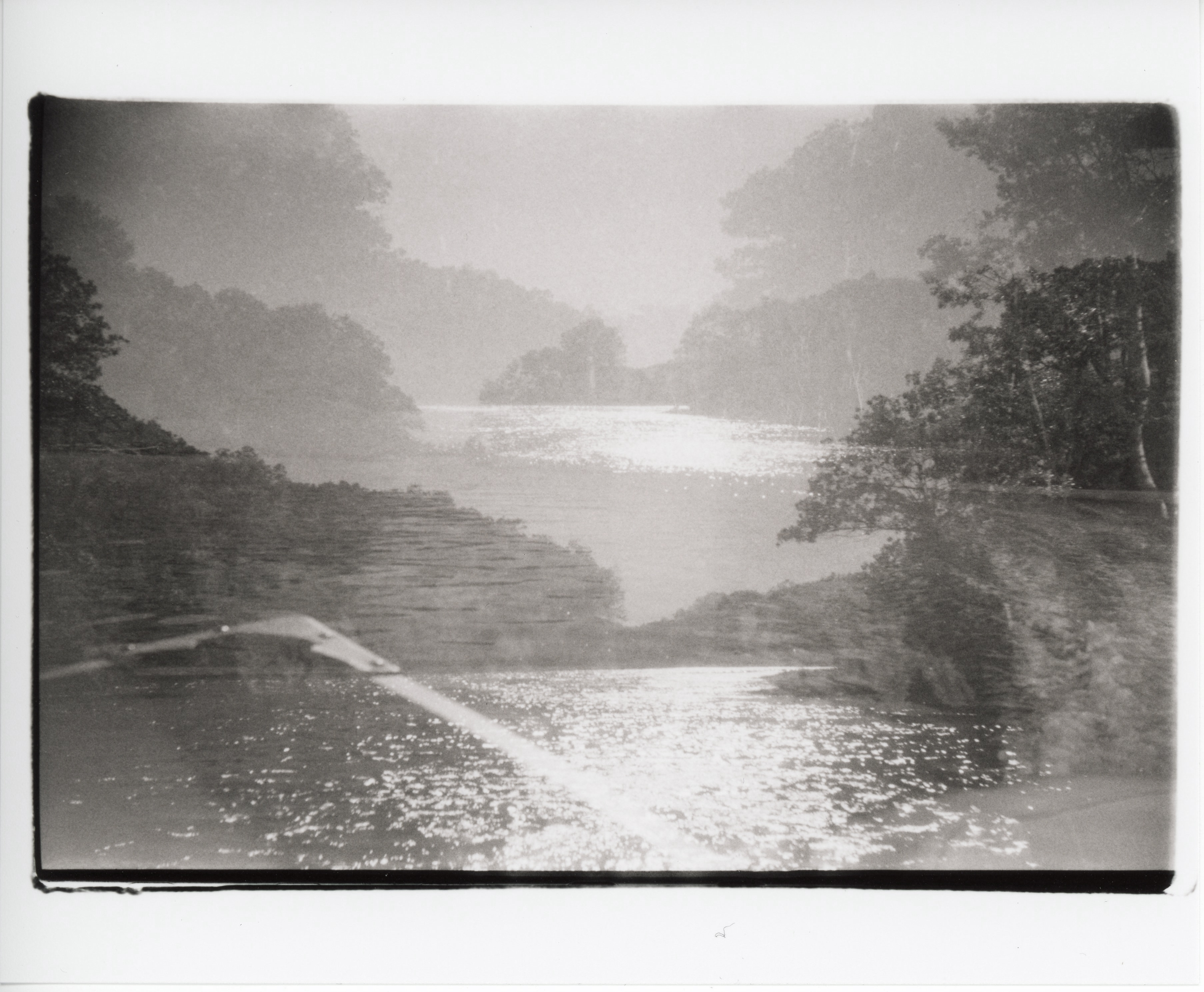 a black and white, sepia toned, 35mm print with messy edges; three similar images looking down a river from a boat are exposed, with the camera shifting vertically between exposures; the bow of the boat is visible in the bottom left corner, only in one of the exposures. there are trees in the distance and trees on either side; the sun is shining on the water