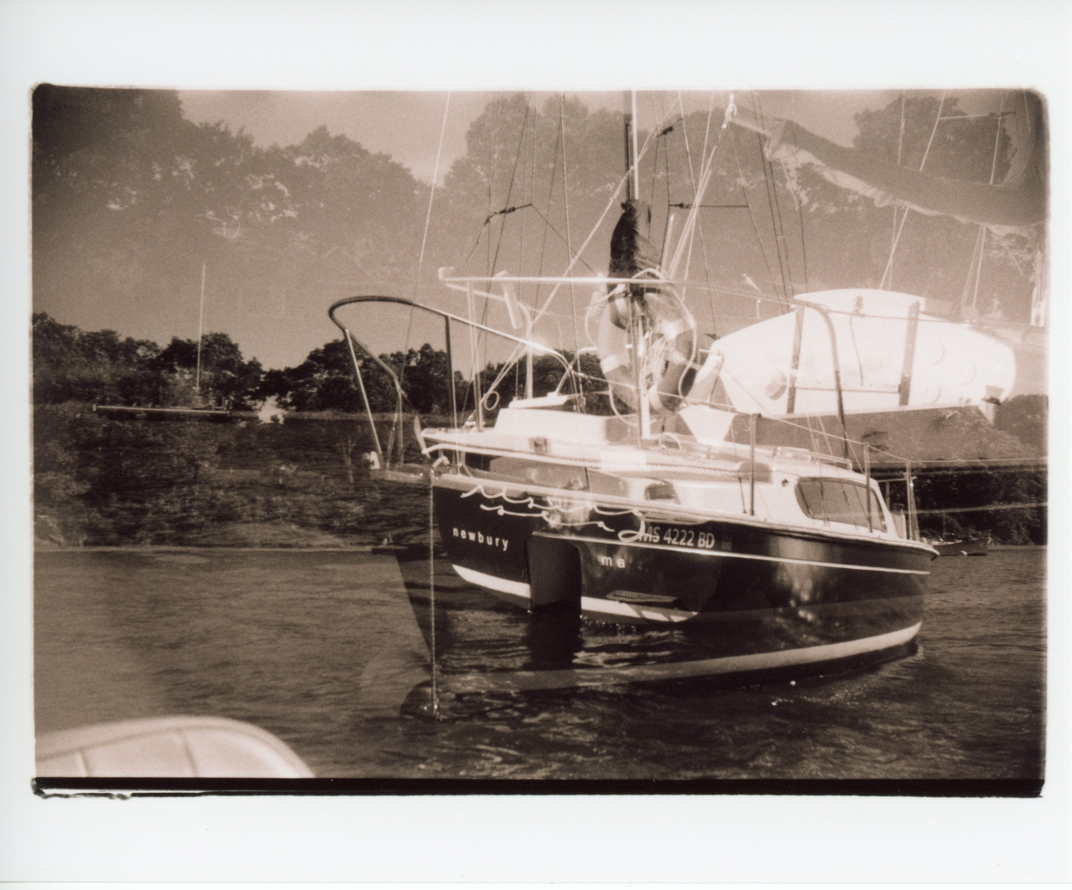 a black and white, sepia toned, 35mm print with messy edges; the image is double exposure of the bow and stern of a boat; on the transom in white cursive letters it says rising sea below in serif lowercase: newbury, ma, MS 4222 BD; there is a strip of land and trees in the background
