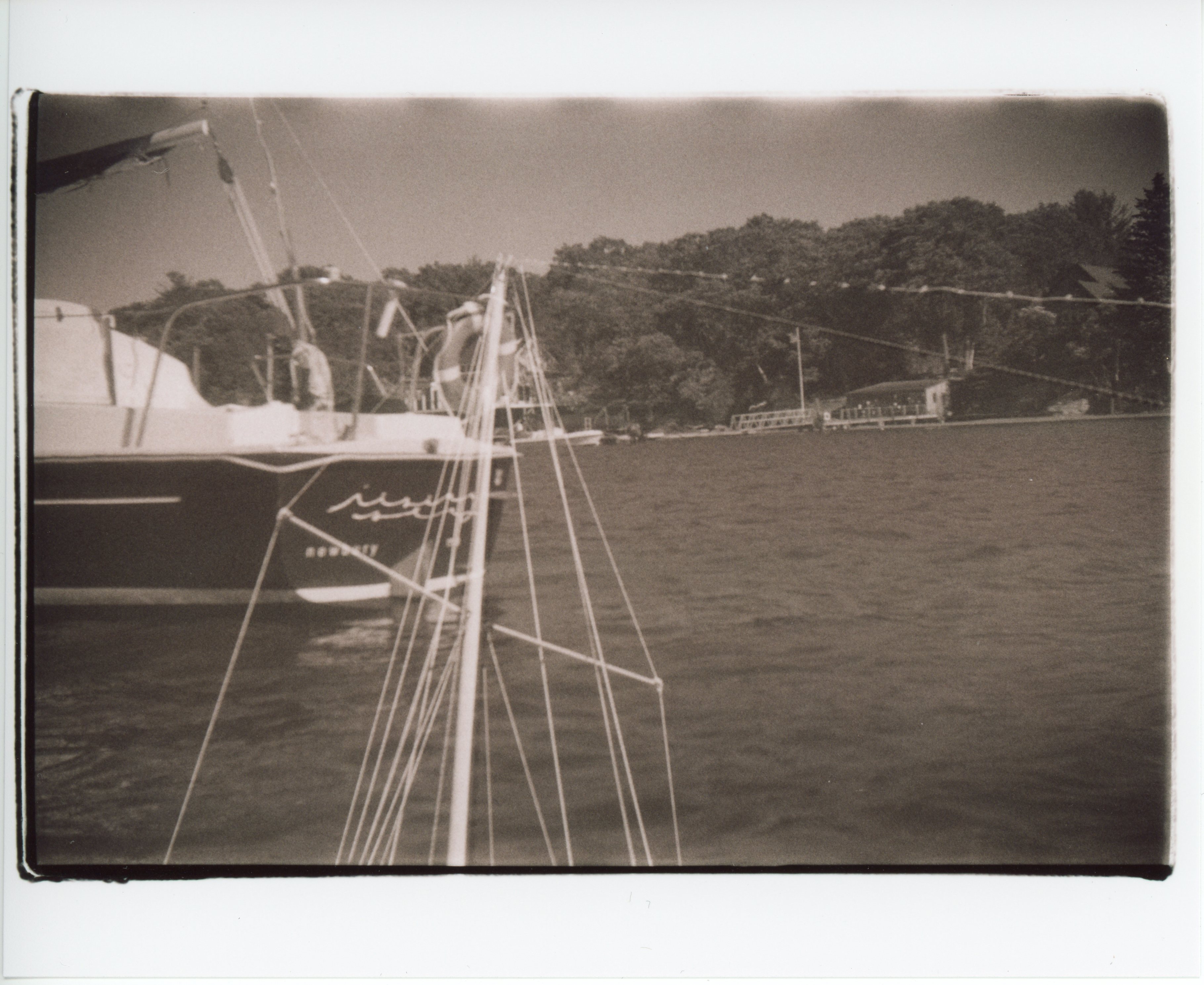 a black and white, sepia toned, 35mm print with messy edges; the image is double exposure of the mast and lines against a clear sky overlaid on an image of the stern of a boat in a harbor; on the transom in white cursive letters it says rising sea, below in serif lowercase: newbury, ma; there is a strip of land and trees in the background