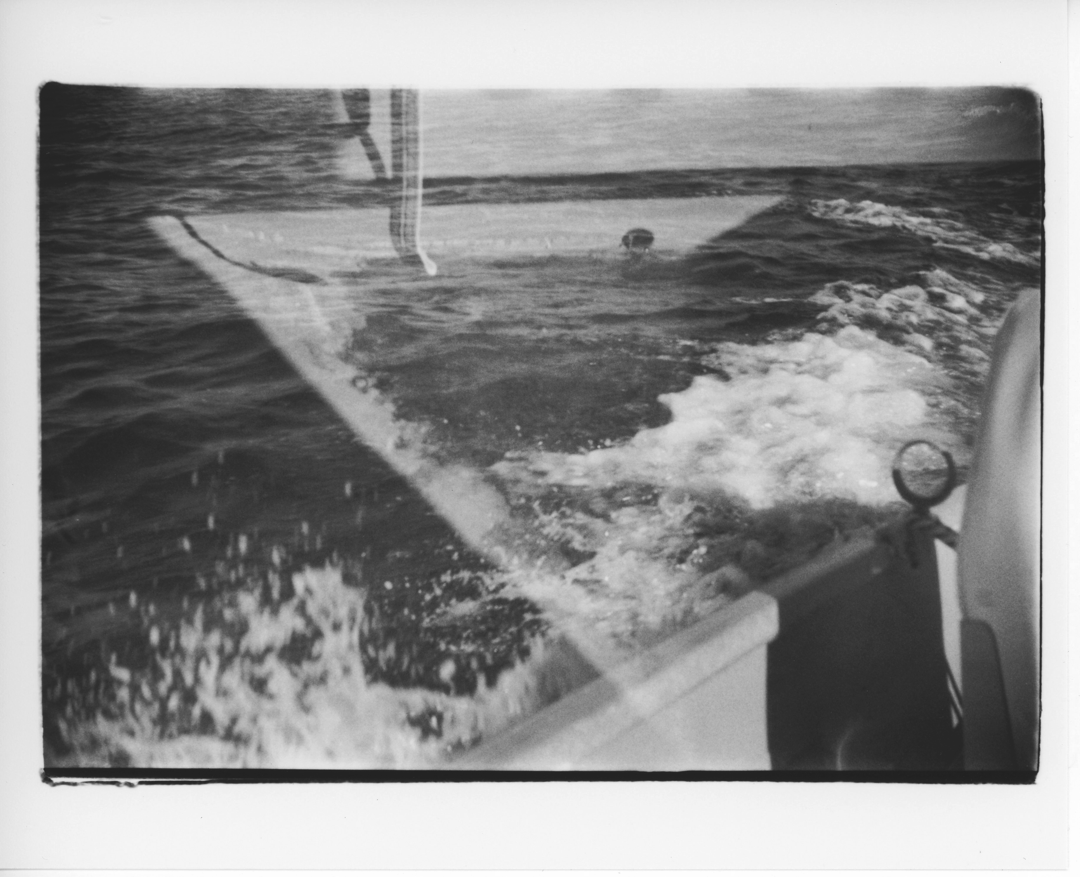 a black and white, 35mm print with messy edges; the image is a double exposure shot from inside a small aluminum outboard motor boat; one image shows the oar rest and the side of the boat in the bottom right corner and the rest of the frame is foam and water next to the boat; the other image shows the sump and the light and shadows in the bottom of the boat