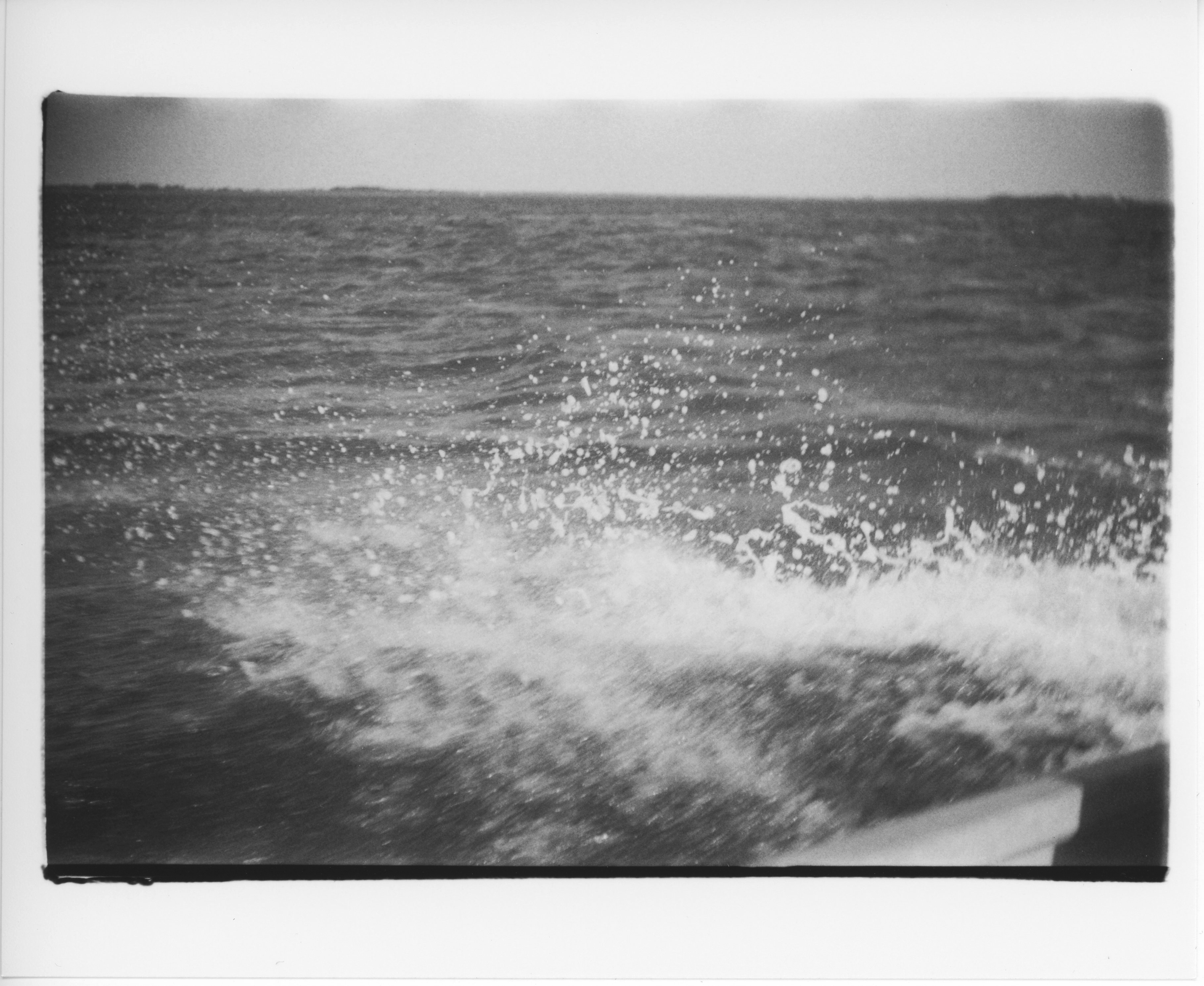 a black and white, 35mm print with messy edges; the photo is shot from inside a small aluminum outboard motor boat; one image shows the oar rest and the side of the boat in the bottom right corner and the rest of the frame is foam and water next to the boat except for a small strip of clear sky at the top of the frame; the foam spray is splashing up out of the water and is sharply focused against the blurry dark waves in the background