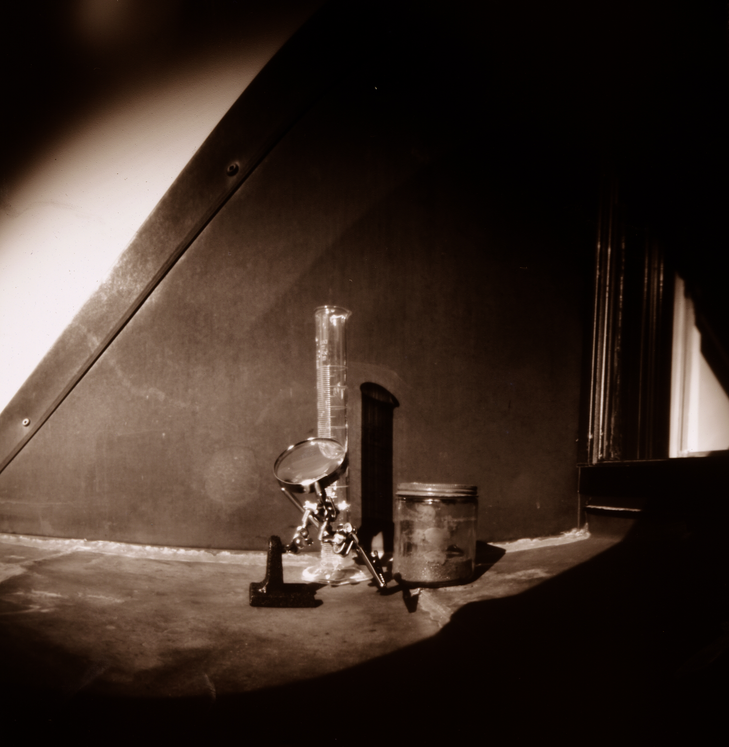a sepia-tinted black and white pinhole camera photograph with a curvature distortion around the edges; the photo is a still life showing an elongated cylinder, an unmarked jar filled with powder, and a magnifying glass on an adjustable stand; the items are arranged in the window well of a window set in the side-roof of a brownstone