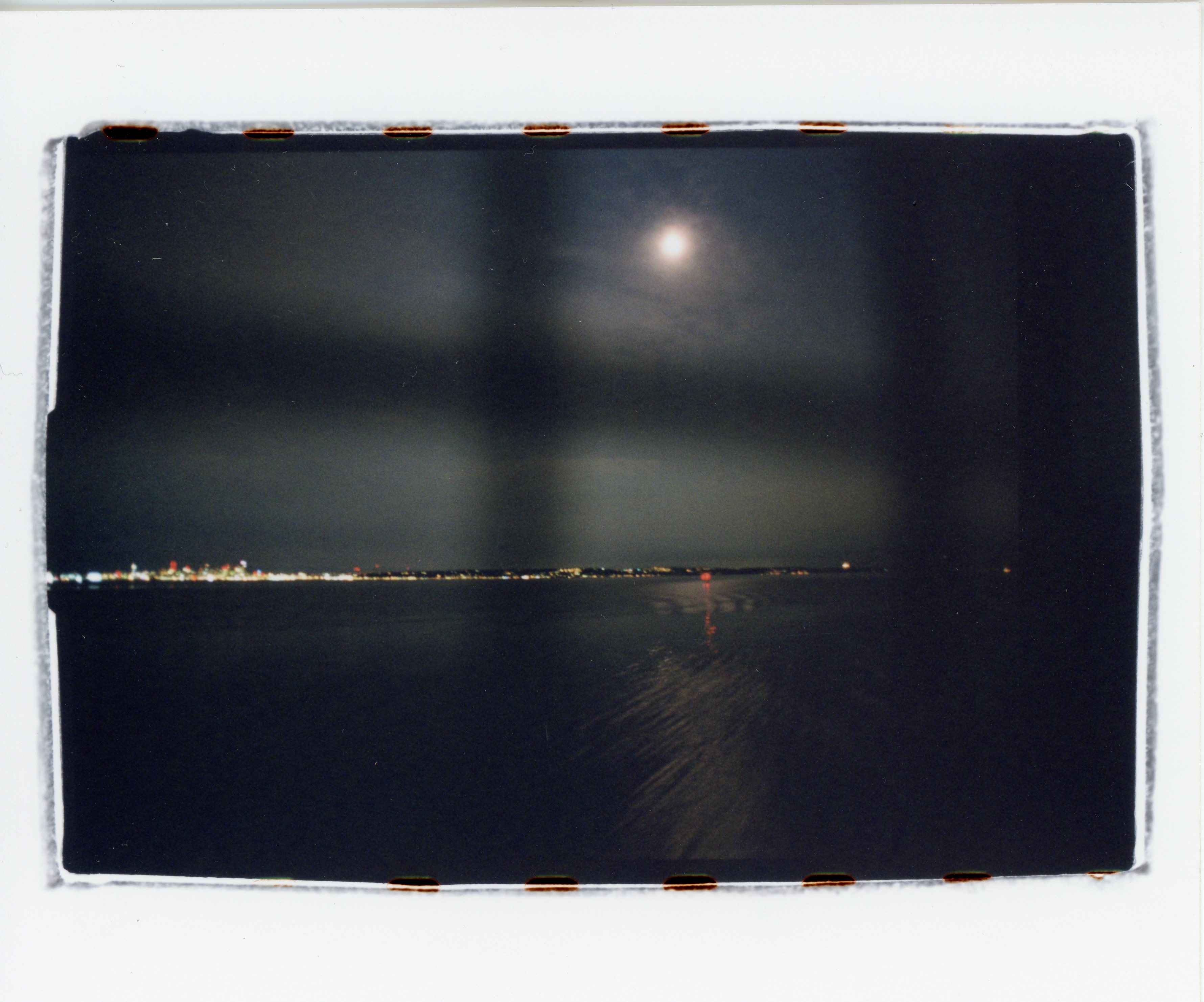 a 35mm color print with messy edges that show bits of the sprocket holes; out of focus directly in front of the camera is a grid mesh on the railings of a ferry; the sky is overcast, but the moon is visible; the sky takes up slightly more than half the top of the frame; there is a thin strip of land between the sky and the water populated by dense lights on the left that dim and disappear on the right side; the moon shines off the waves in the water, which curve towards the land